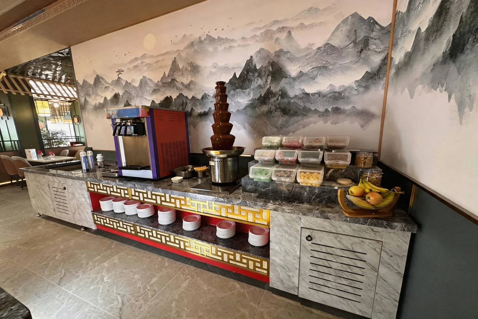 Chocolate fountain and ice cream machine on the buffet counter, in front of a Chinese ink-wash mountain mural